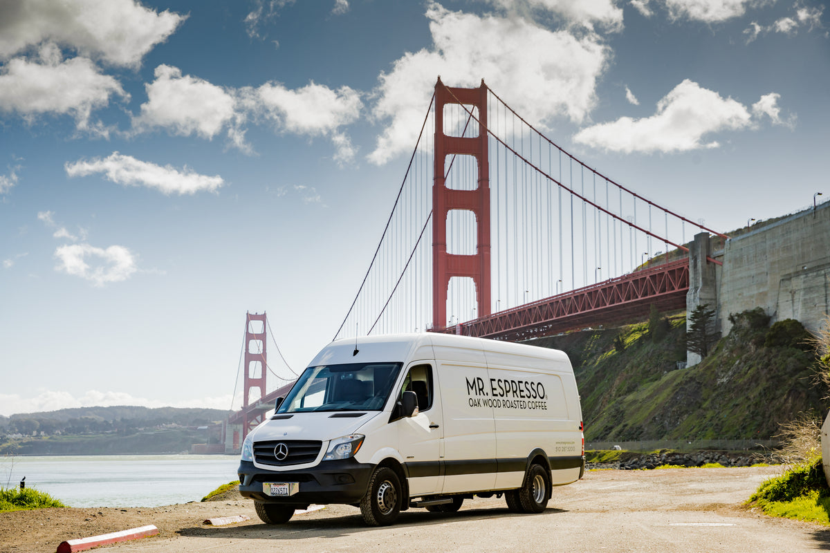 A white Mercedes-Benz Sprinter van with 'MR. ESPRESSO SAN FRANCISCO RESTAURANT' printed on the side, parked outdoors in front of the Golden Gate Bridge under a blue sky with clouds.