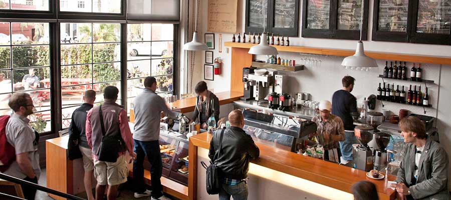 A busy cafe interior with multiple people standing at the counter and ordering, with a wood counter, coffee equipment, shelves of bottles, and large windows in the background.