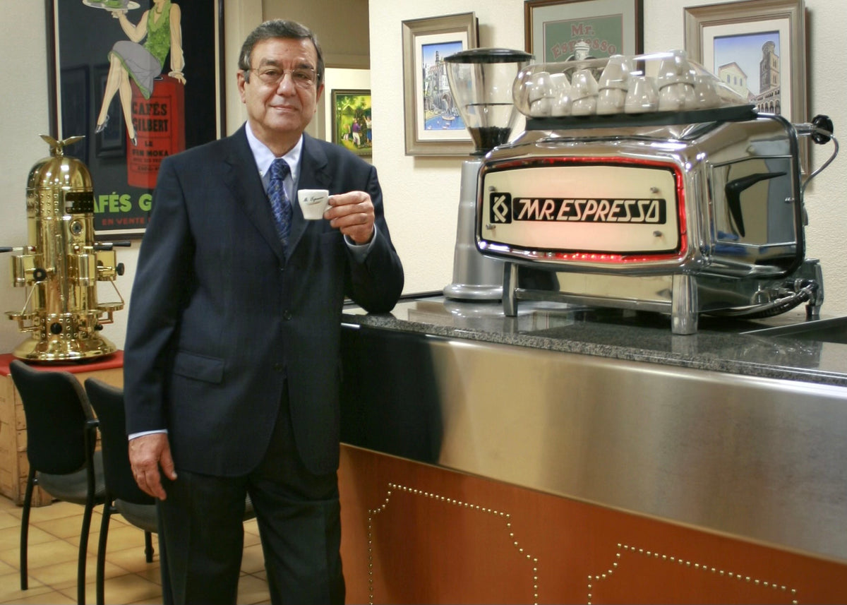 A man in a dark suit holding a small espresso cup stands beside a stainless steel bar counter with a Mar. Espresso machine in a cafe setting.