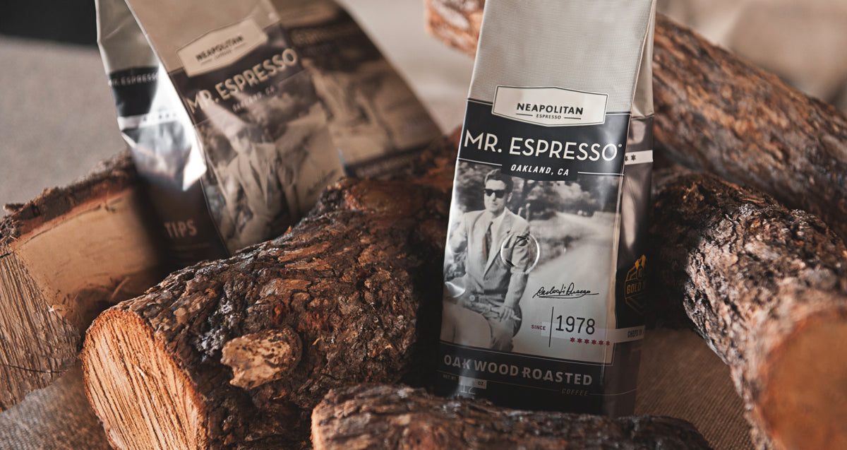 Two bags of Mr. Espresso Oak Wood Roasted coffee beans are displayed on a burlap surface surrounded by rustic wood logs.