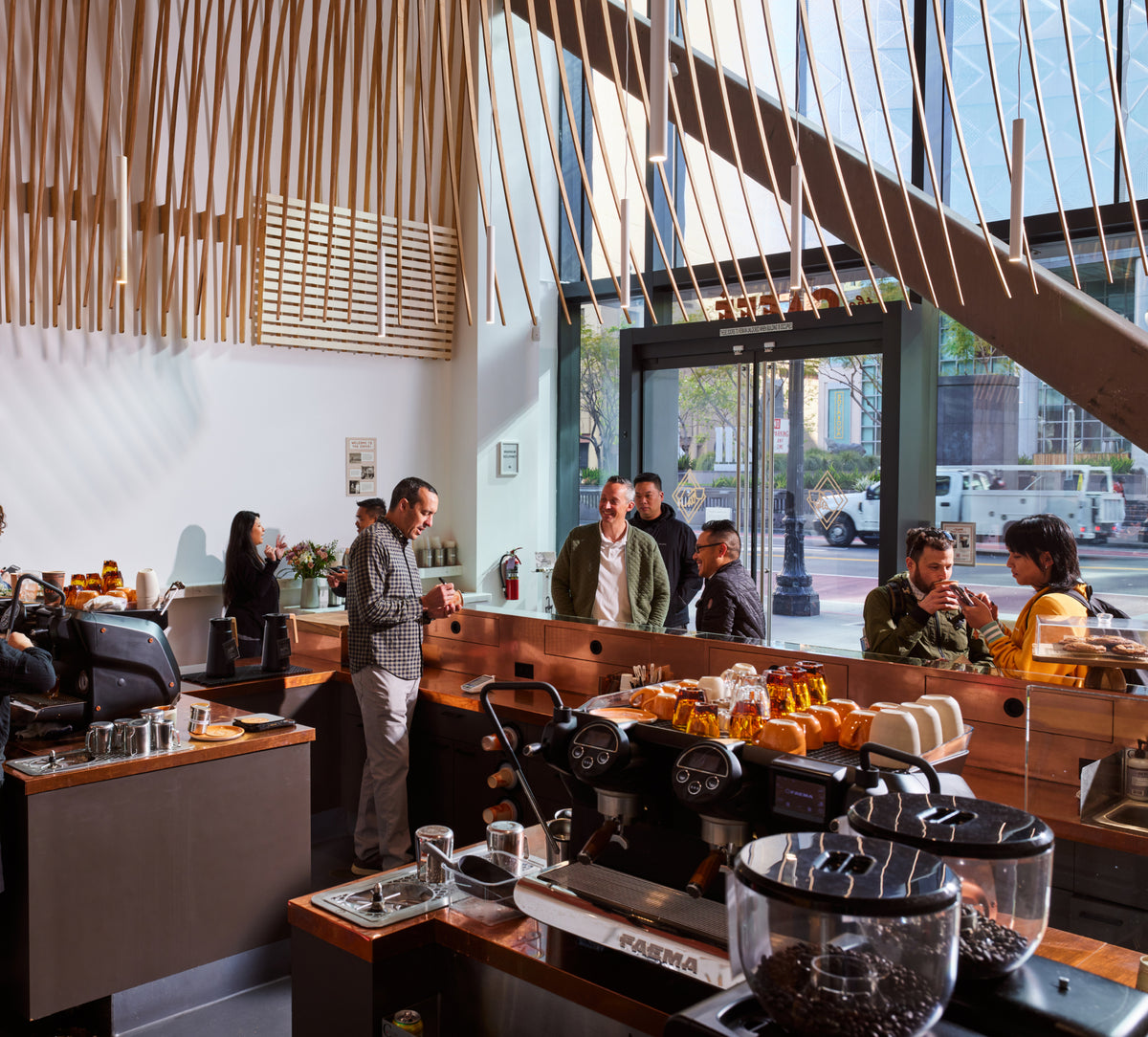 Customers gather inside a bright, modern coffee shop as baristas work behind the counter, with espresso machines, coffee grinders, and warm wood details throughout the space.