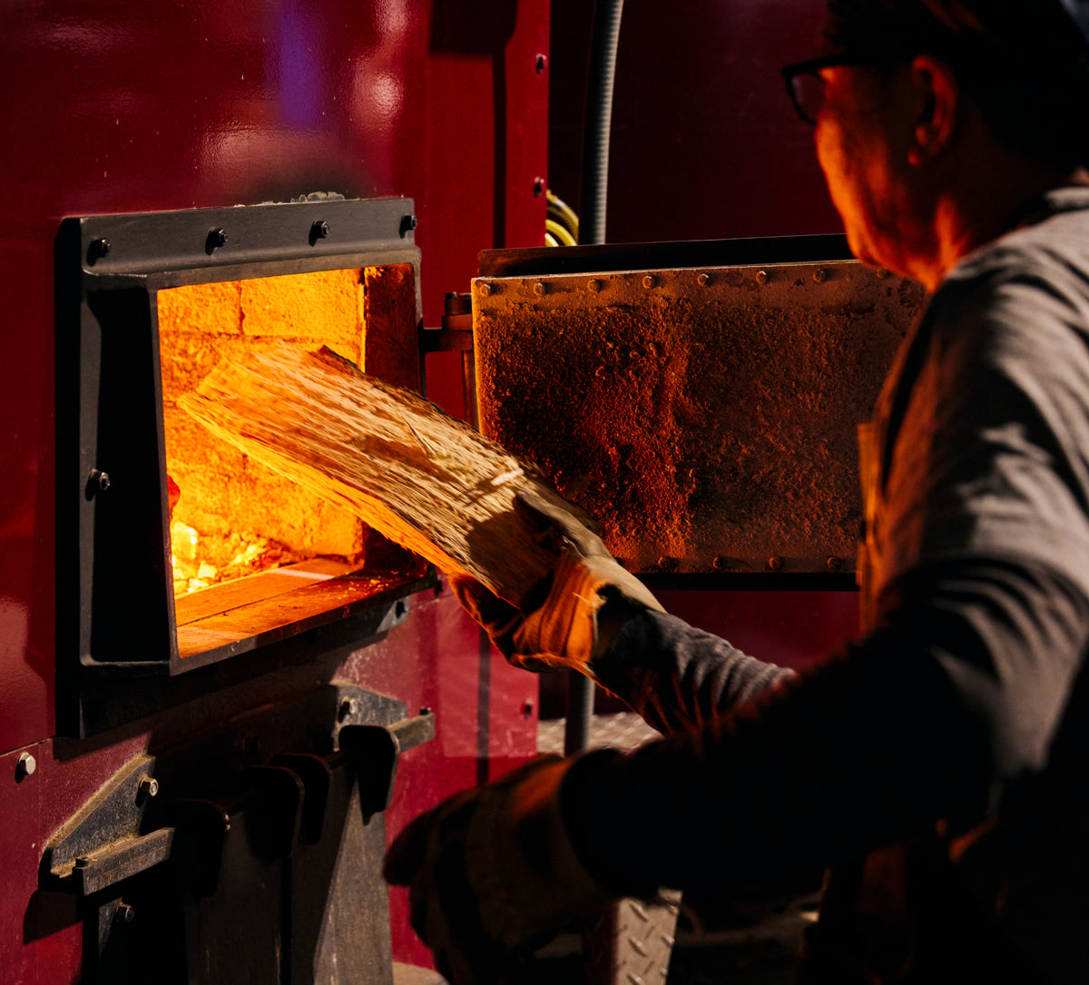 A worker wearing protective gloves feeds wooden boards into a glowing industrial furnace, illuminated by the orange heat of the fire.