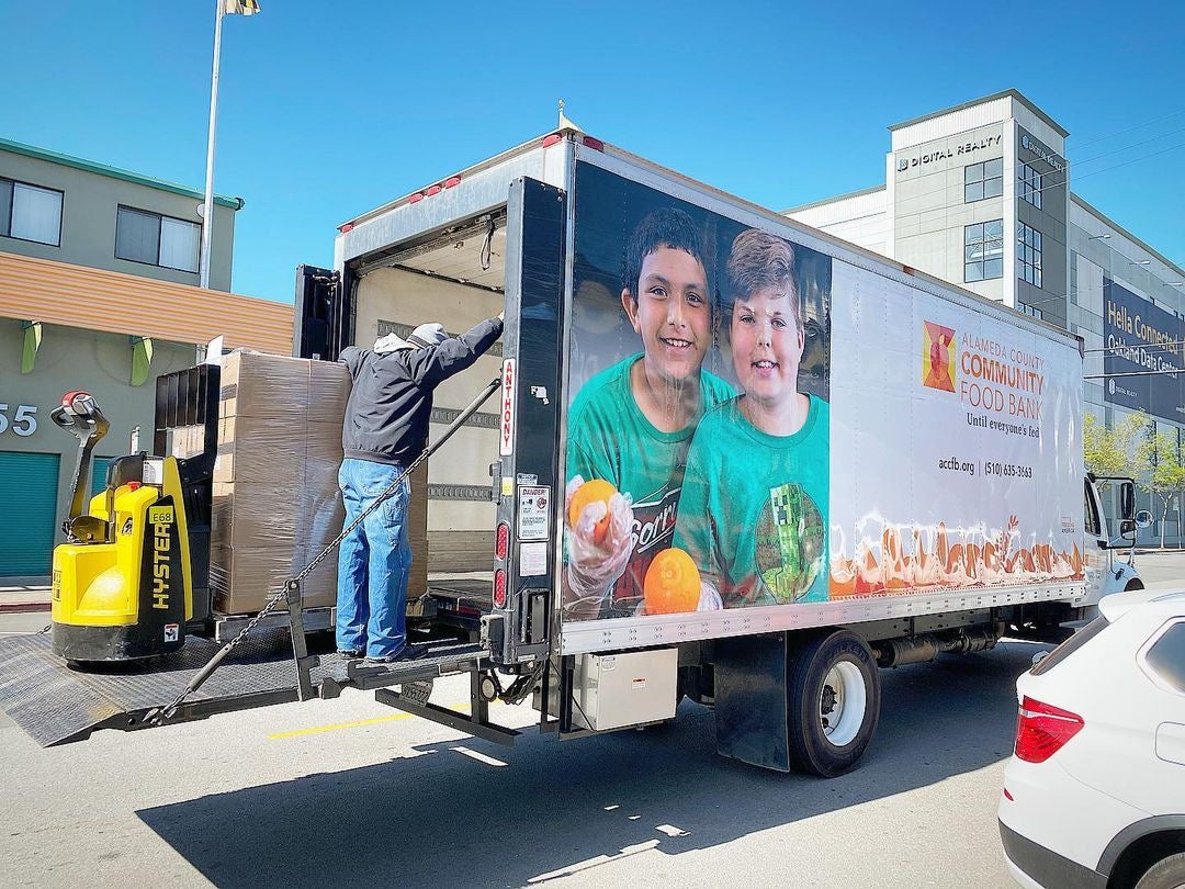 A white delivery truck parked on a street with its rear doors open, showing a large printed advertisement on the side featuring two smiling children and food imagery. A worker is standing inside the truck near the cargo area, and a yellow Hyundai forklift is visible on the left.