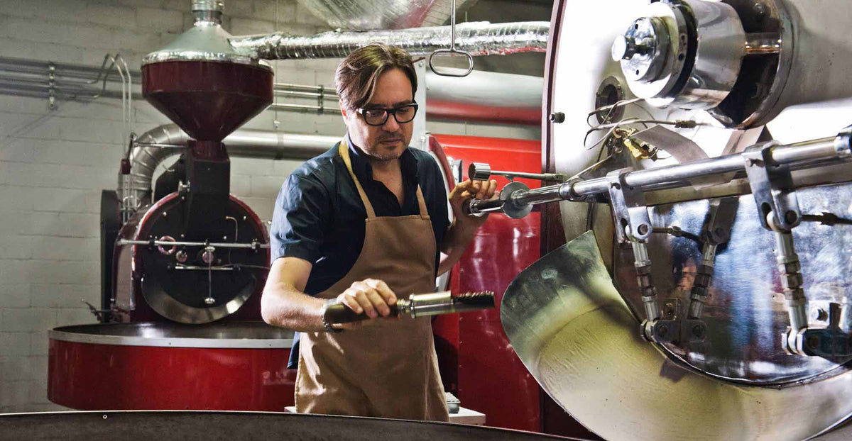 A man wearing glasses, a dark blue shirt, and a tan apron operates a large stainless steel coffee roaster in a coffee roastery.