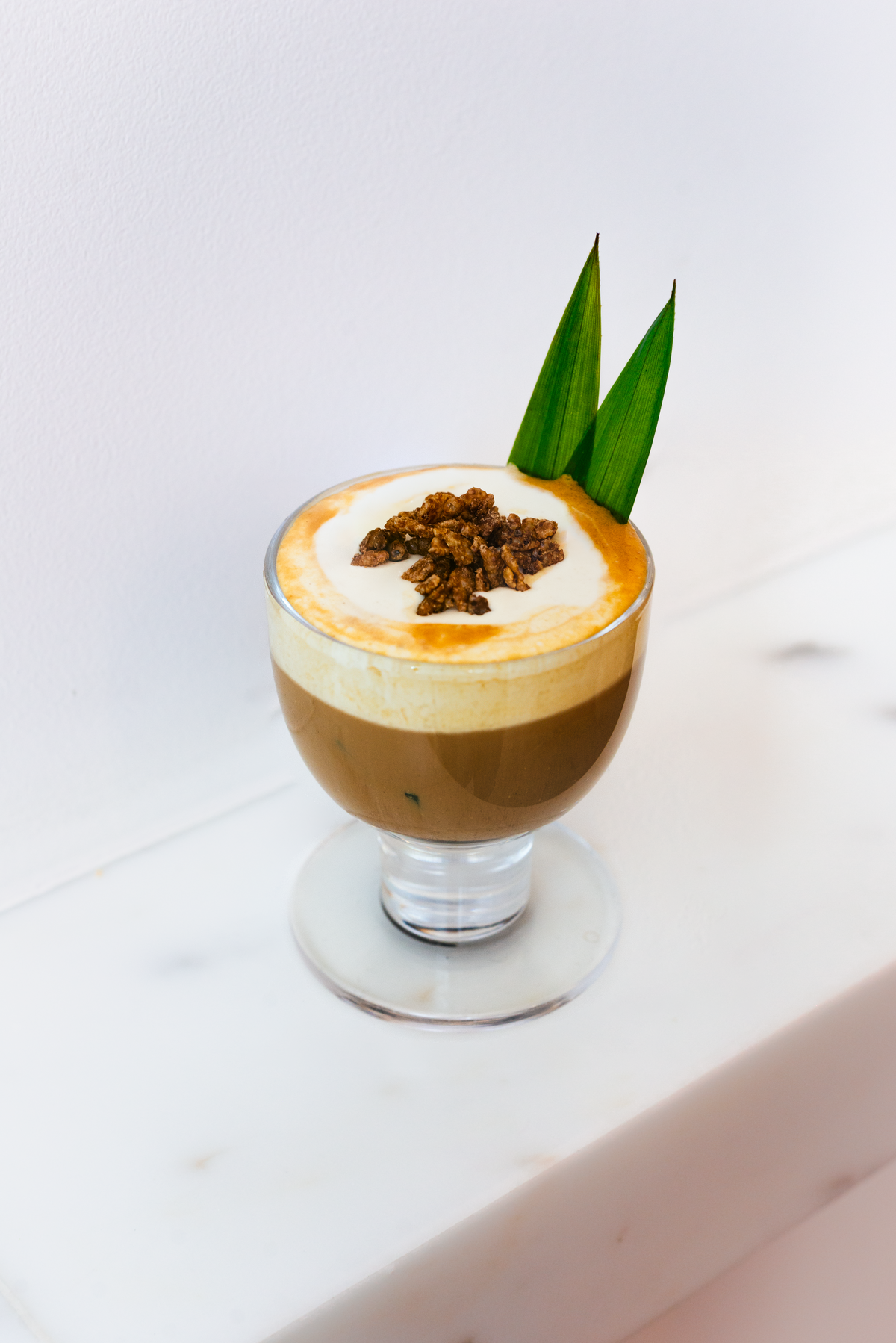 A layered coffee drink in a clear glass cup, topped with whipped cream, a sprinkle of brown granola, and two green leaf garnishes, set on a white marble surface against a white wall.