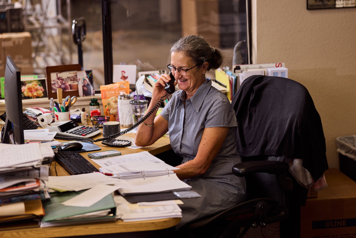 A woman with gray hair tied back sits at a cluttered desk, smiling while talking on a landline telephone. The office is filled with papers, files, a computer monitor, and office supplies.