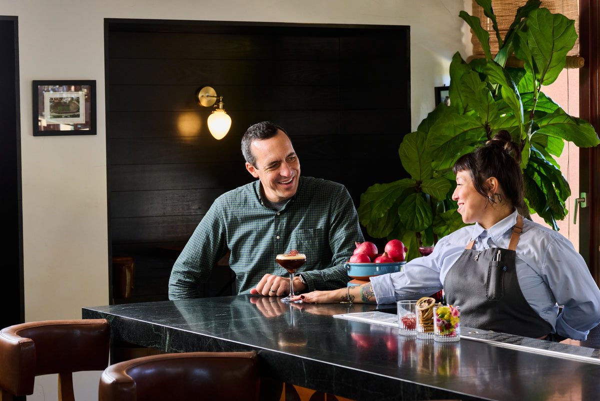 A man and a woman converse at a dark bar counter in a restaurant or cafe. The man wears a dark green textured shirt and holds a cocktail glass, while the woman wears a gray apron over a light blue shirt.