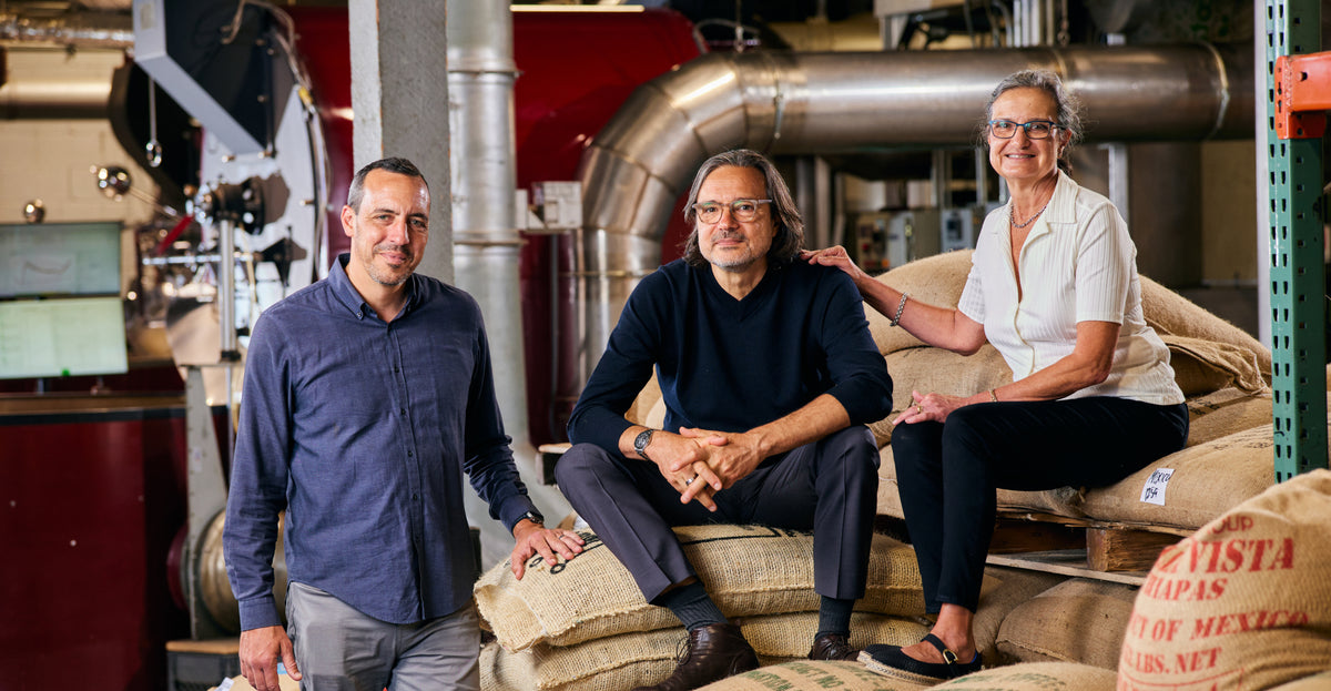 Three people standing and sitting among large burlap sacks in a rustic industrial setting with metal pipes and machinery in the background.