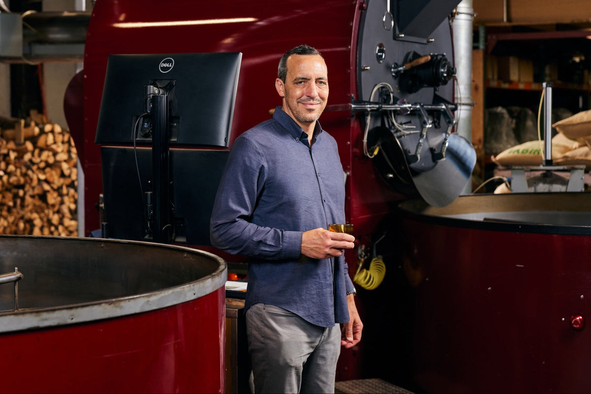 A man in a blue button-down shirt and gray pants stands in front of a large red coffee roaster inside a roastery, holding a small glass of coffee.