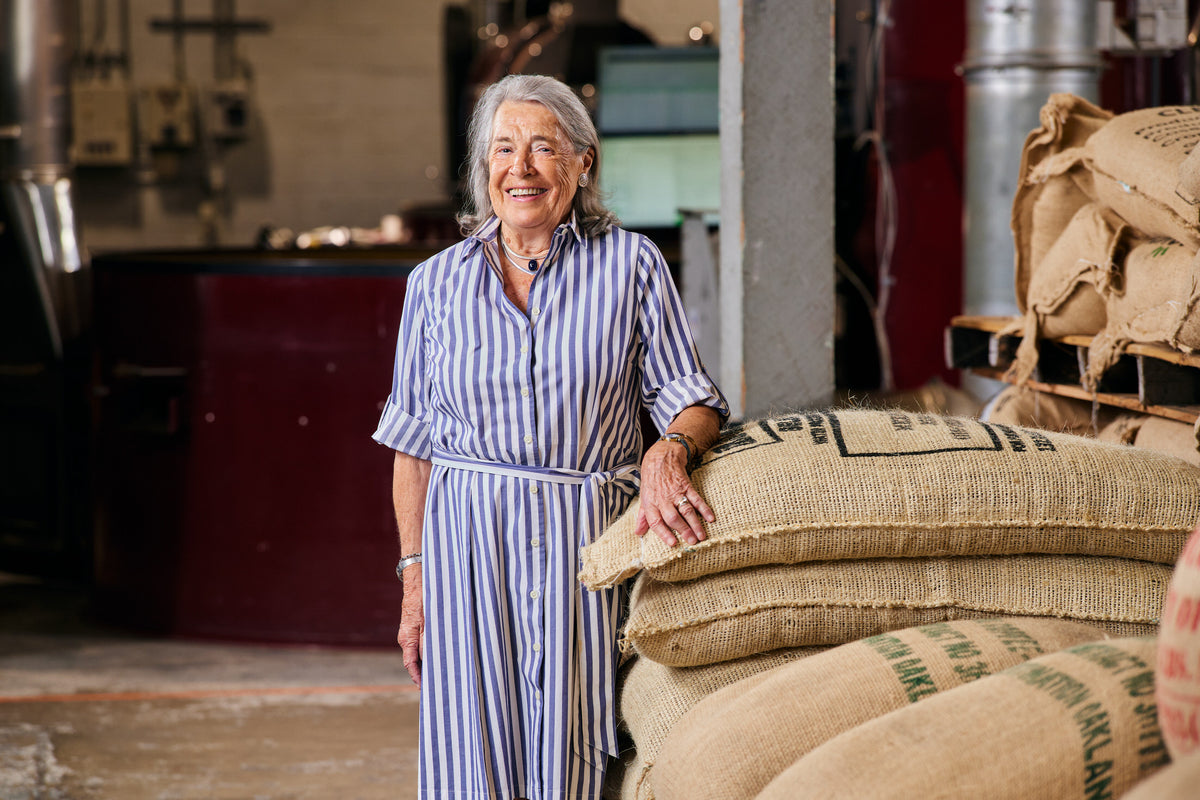 A smiling woman with gray hair stands in a rustic indoor setting, wearing a blue and white vertically striped button-down shirt dress. She is leaning on a large burlap sack, with more sacks stacked behind her and a warm-toned interior background.