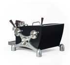 A modern espresso machine made by Slayer with a matte black body, polished silver metal legs and valves, and dark brown wooden handles, shown on a plain white background.