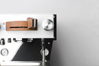 Close-up of a modern espresso machine with a white top panel, brushed silver metal body, wood-grain handle detail, and black structural elements against a light gray background.