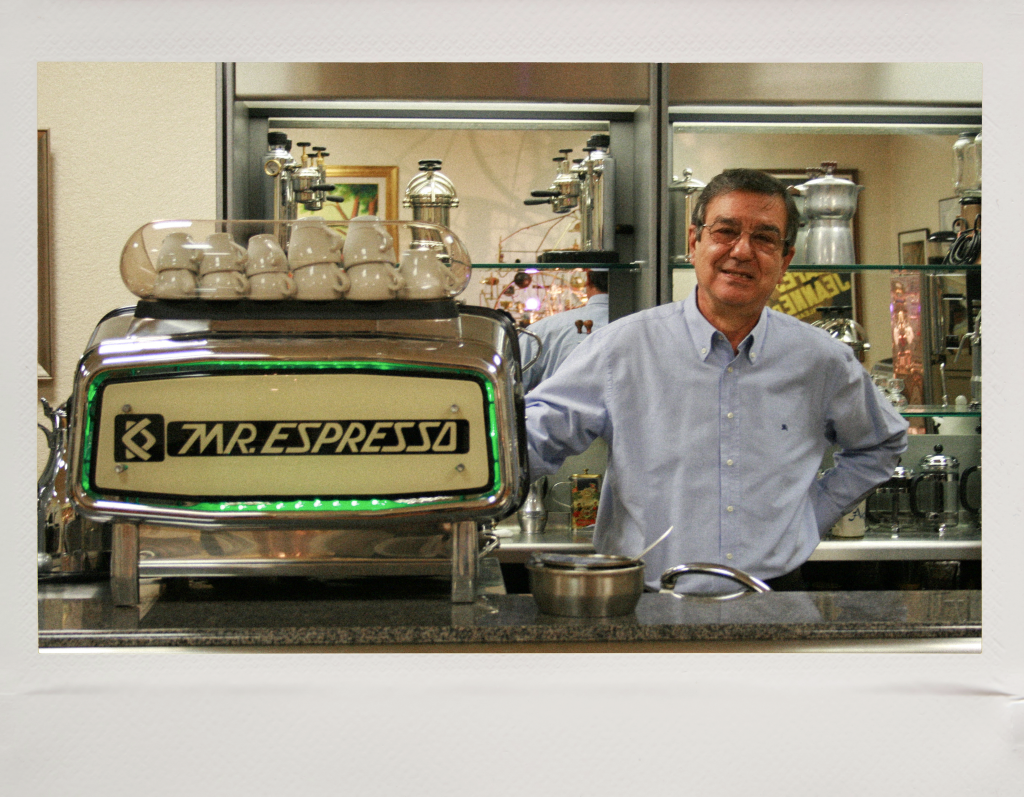 Carlo Di Ruocco in a light blue button-down shirt standing behind a Mr. Espresso machine, with coffee cups and equipment visible in the background.