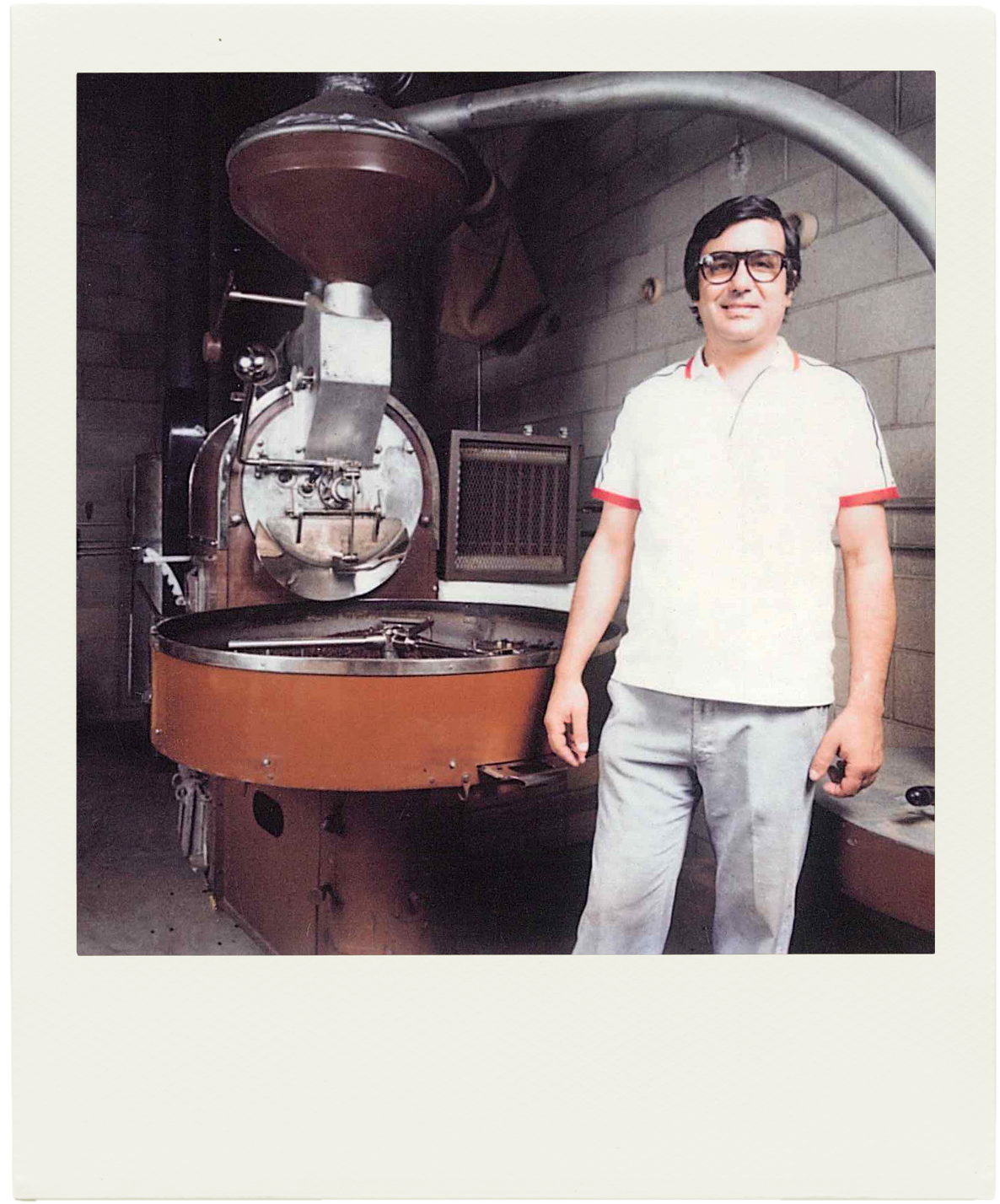 Founder Carlo Di Ruocci in black glasses, a white polo shirt with red trim, and light gray pants standing next to a large coffee roasting machine. The photo has a vintage Polaroid-style border.