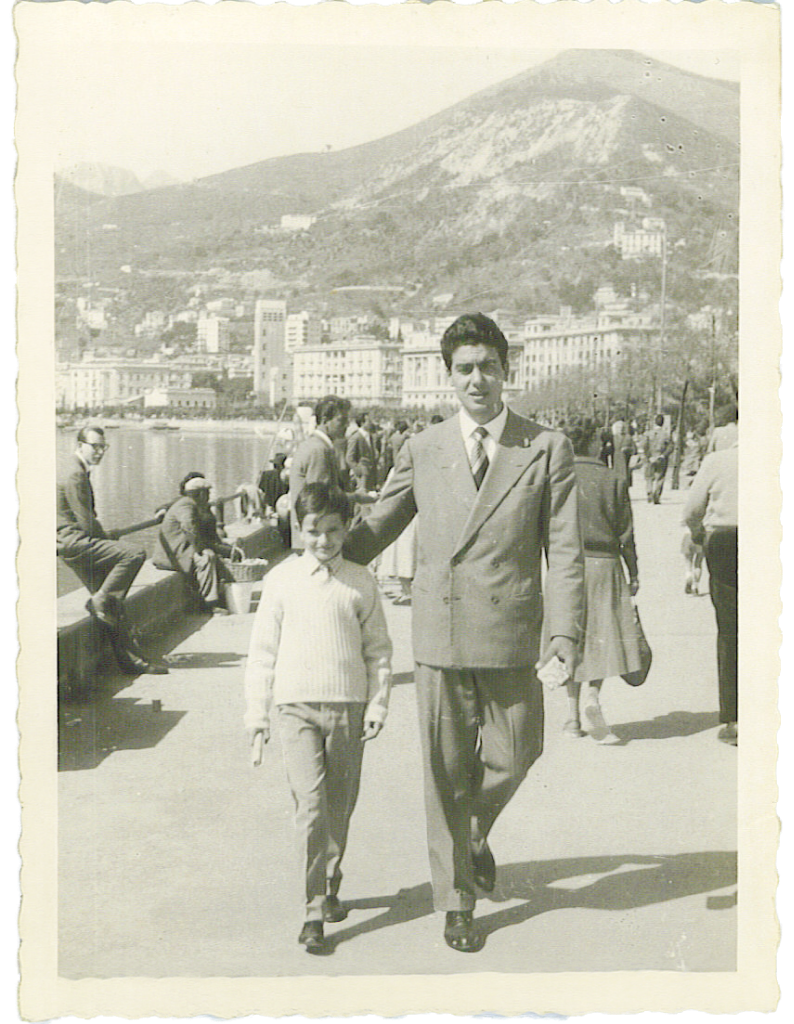 Black and white vintage photograph of a man in a suit walking arm-in-arm with a Carlo Di Ruocco as young boy in Salerno, Italy with buildings and a mountain in the background.