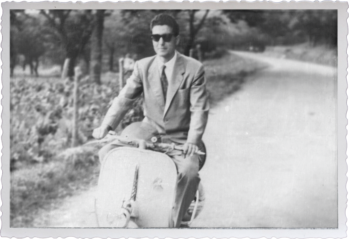 Black and white vintage photograph of a Carlo Di Ruocco in a suit and sunglasses riding a Vespa scooter in Italy. This is Mr. Espresso's signature photo. 