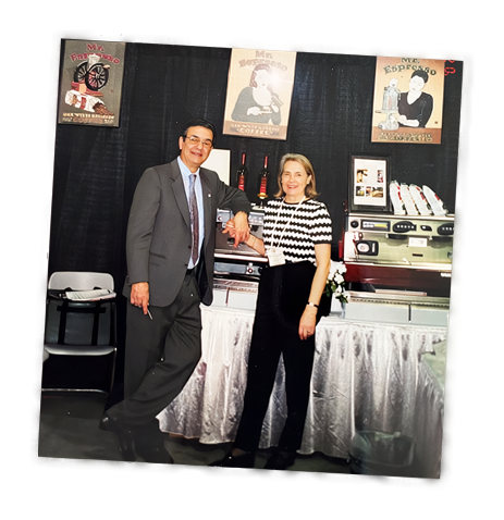 A vintage-style photograph showing Carlo Di Ruocco in a gray suit and wife Marie Di Ruocco in a black-and-white patterned top in front of a display of espresso machines with framed posters on the wall.
